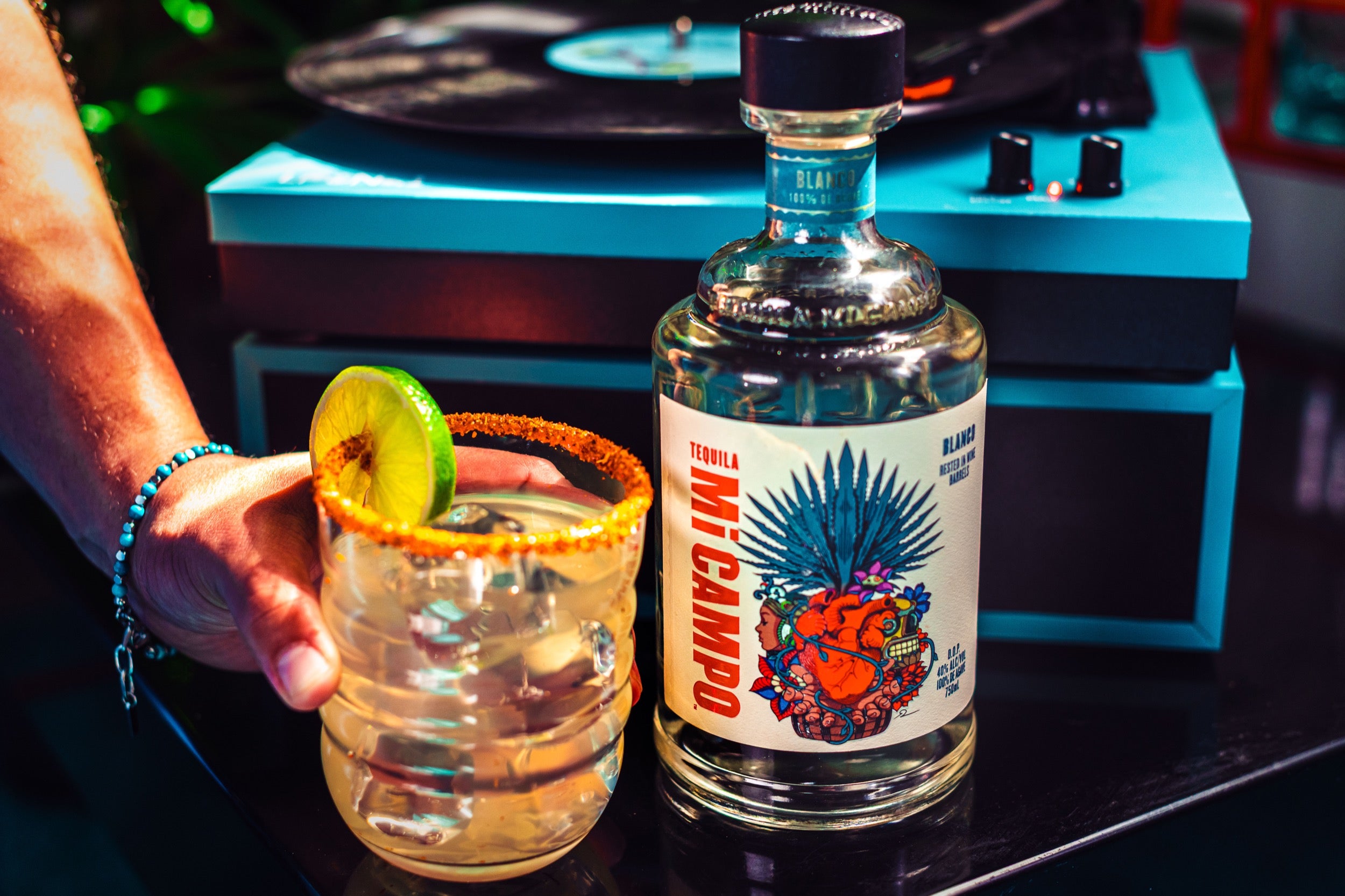 Bottle of Mi Campo with a cocktail garnished with a lime, held by a person in a club setting.