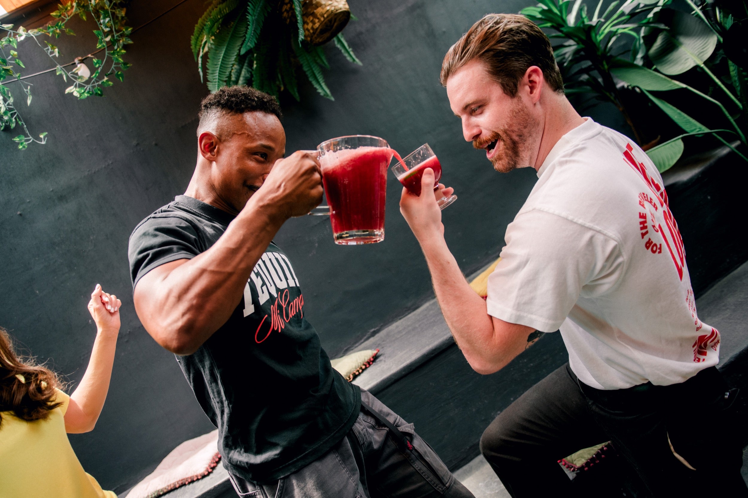 Two men in casual clothing interacting with drinks against a dark background with plants.