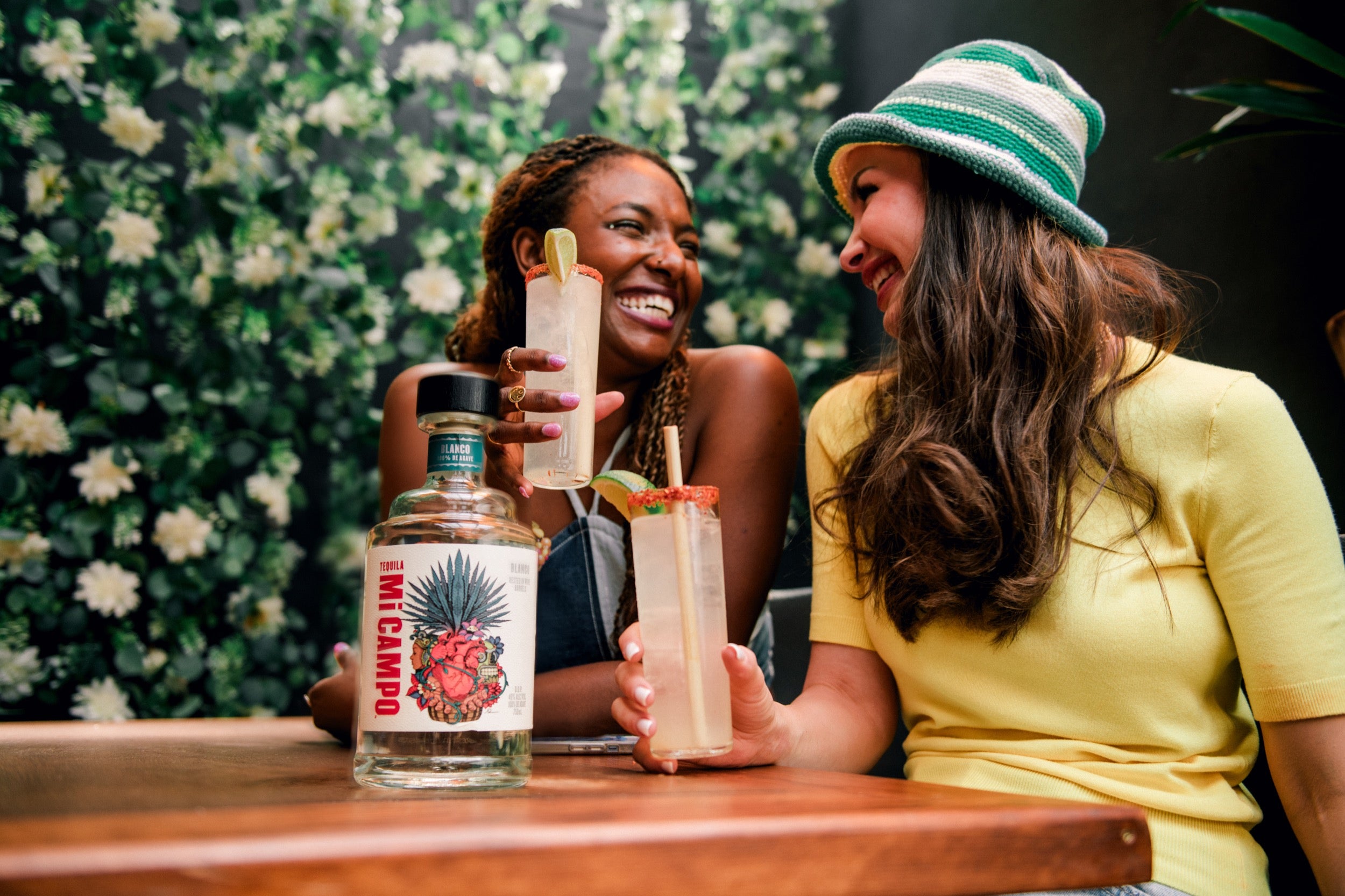 Two people sitting at a table with a bottle of Mi Campo and a cocktail, against a floral wall.