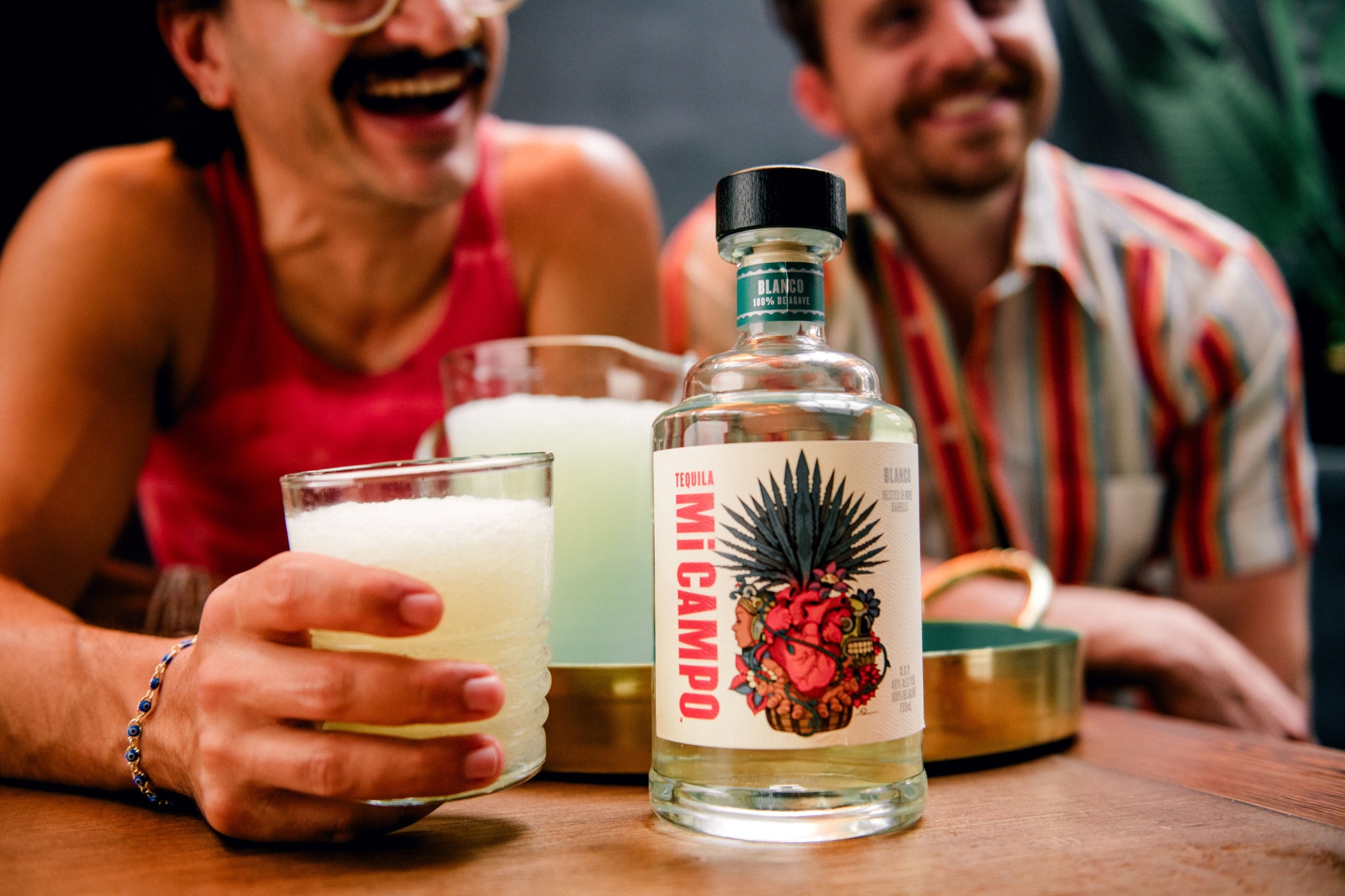 Two people sitting at a table with a bottle of Mi Campo tequila and glasses.