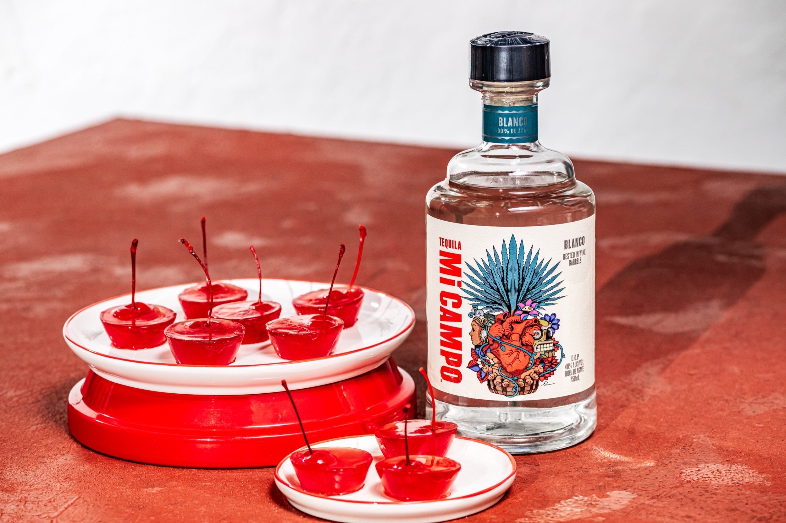 Bottle of Mi Campo Blanco tequila beside small red cherry jelly shots topped with stems, arranged on white plates against a warm terracotta background.