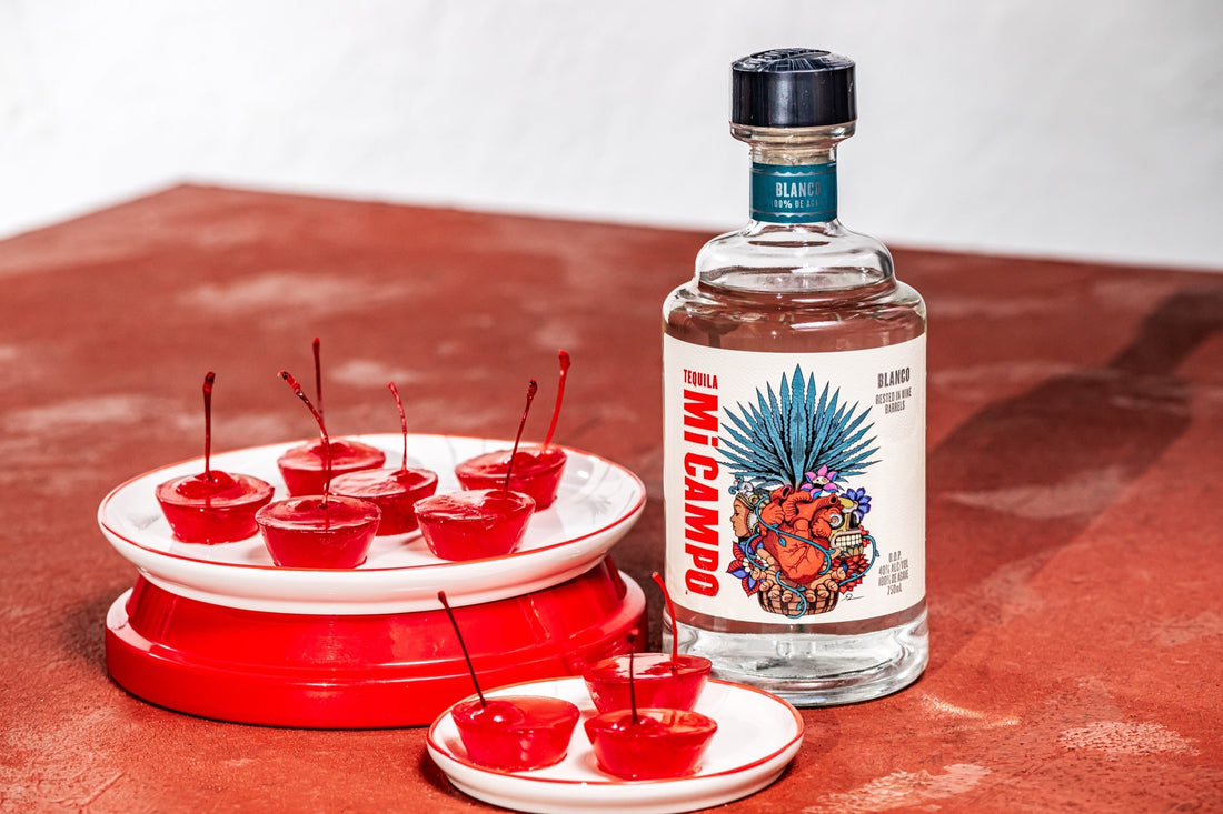Bottle of Mi Campo Blanco tequila beside small red cherry jelly shots topped with stems, arranged on white plates against a warm terracotta background.