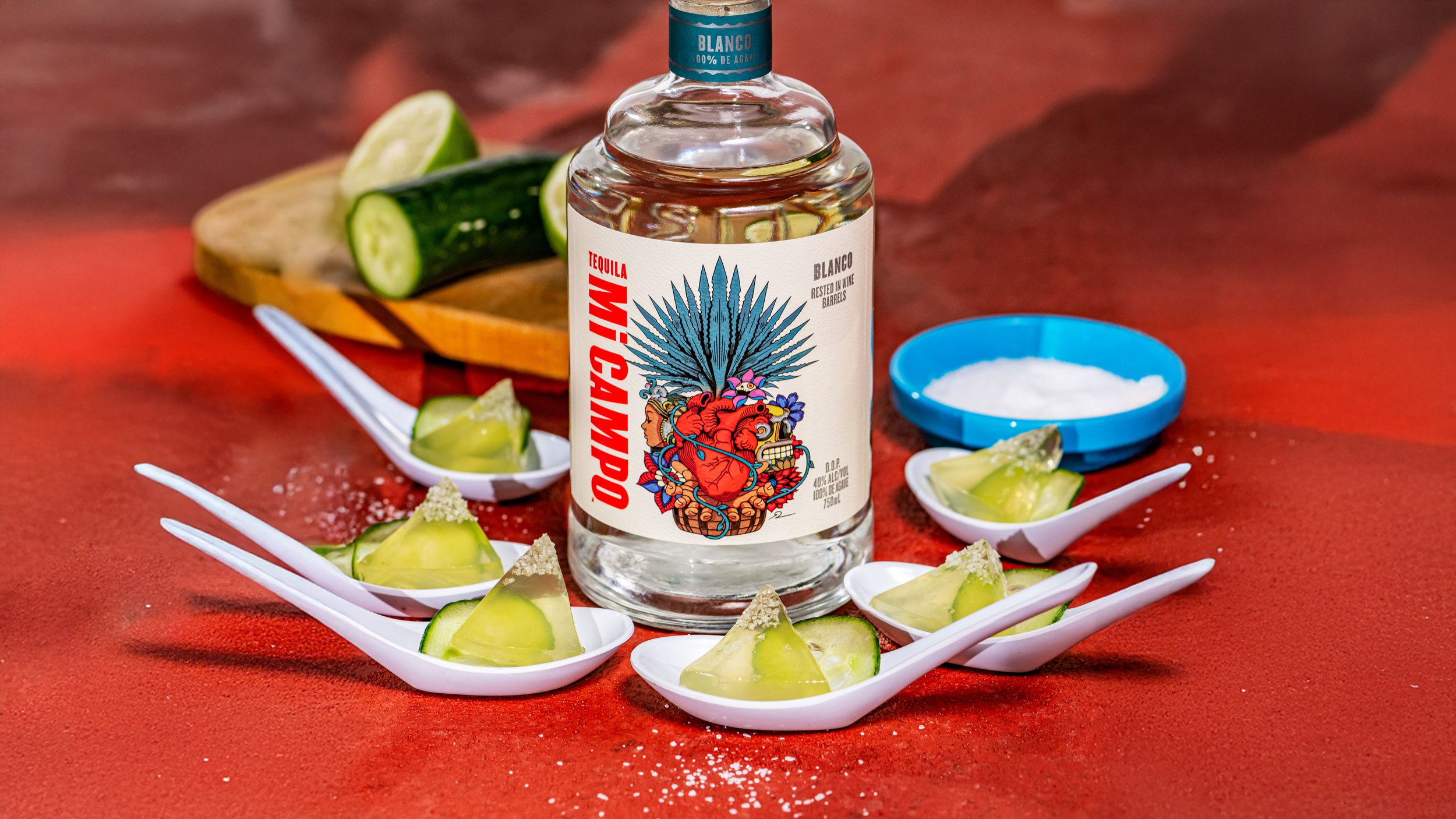 Bottle of Mi Campo Blanco tequila surrounded by cucumber margarita jelly shots served on white tasting spoons with cucumber slices and salt.