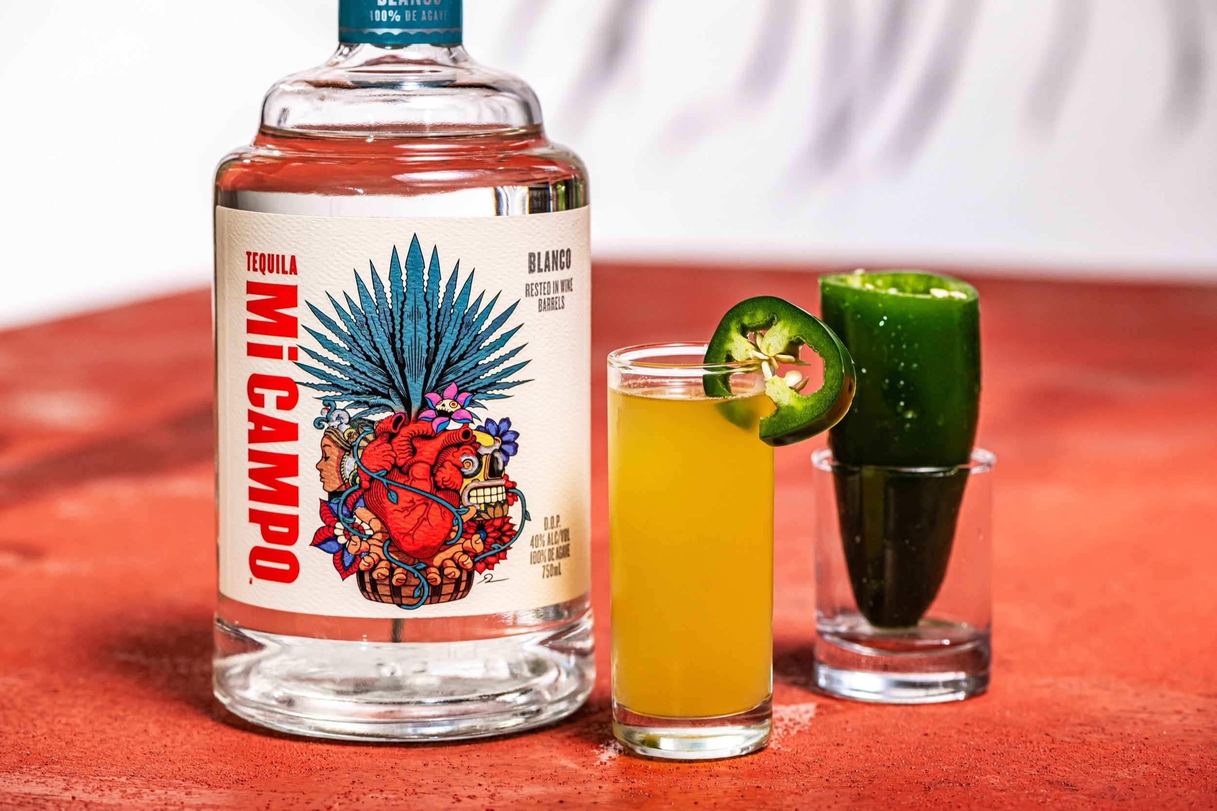 Bottle of Mi Campo Blanco tequila beside a jalapeño popper shooter and a jalapeño pepper shot glass on a terracotta surface.