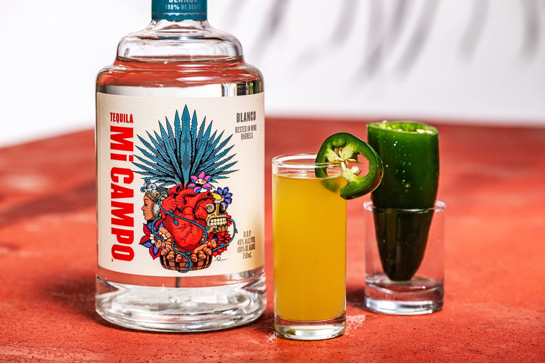 Bottle of Mi Campo Blanco tequila beside a jalapeño popper shooter and a jalapeño pepper shot glass on a terracotta surface.