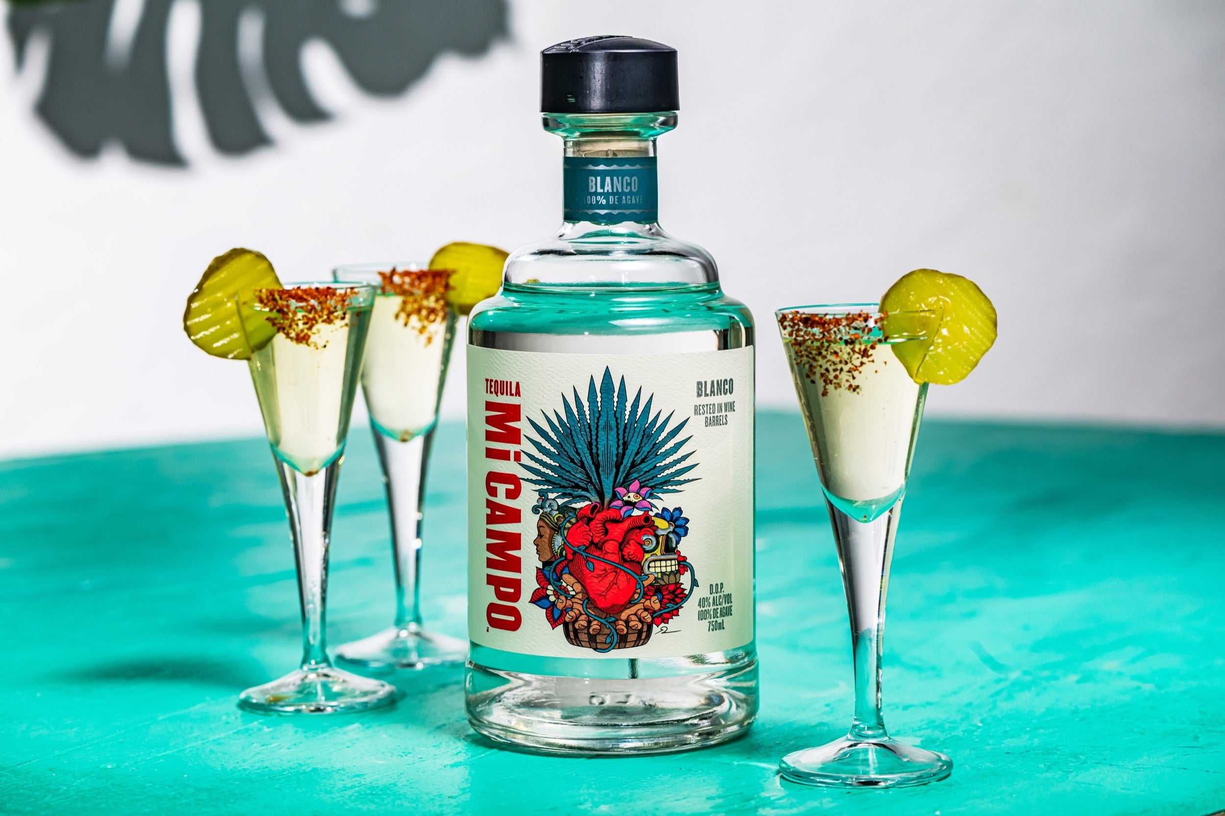 Bottle of Mi Campo Blanco tequila beside pickle shots with Tajín rims and pickle slice garnish, served in tall shot glasses on a teal surface.
