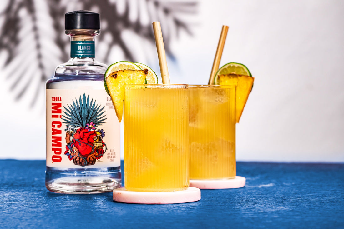 Bottle of Mi Campo Blanco tequila beside two pineapple ginger crush cocktails garnished with pineapple and lime, served over ice with straws.