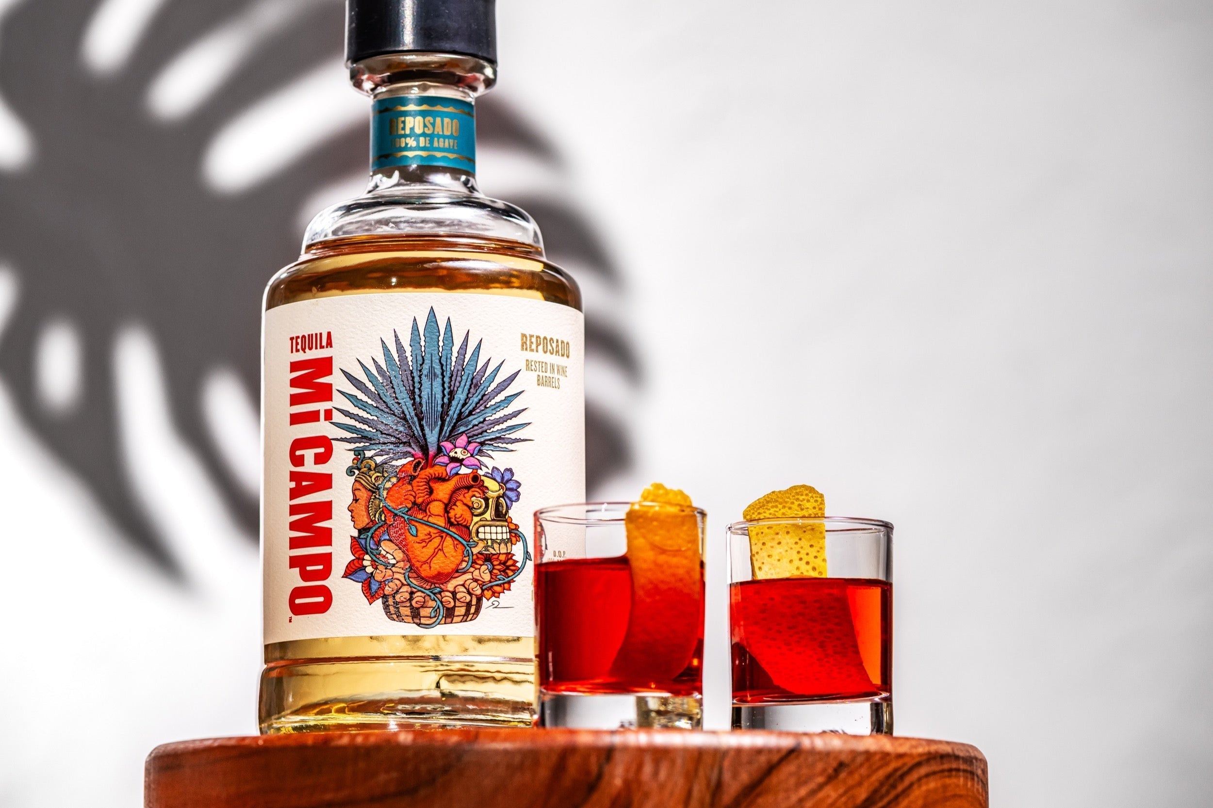Bottle of Mi Campo Reposado tequila beside two red Rosita shooters garnished with orange peel, set on a wooden surface with soft shadows in the background.