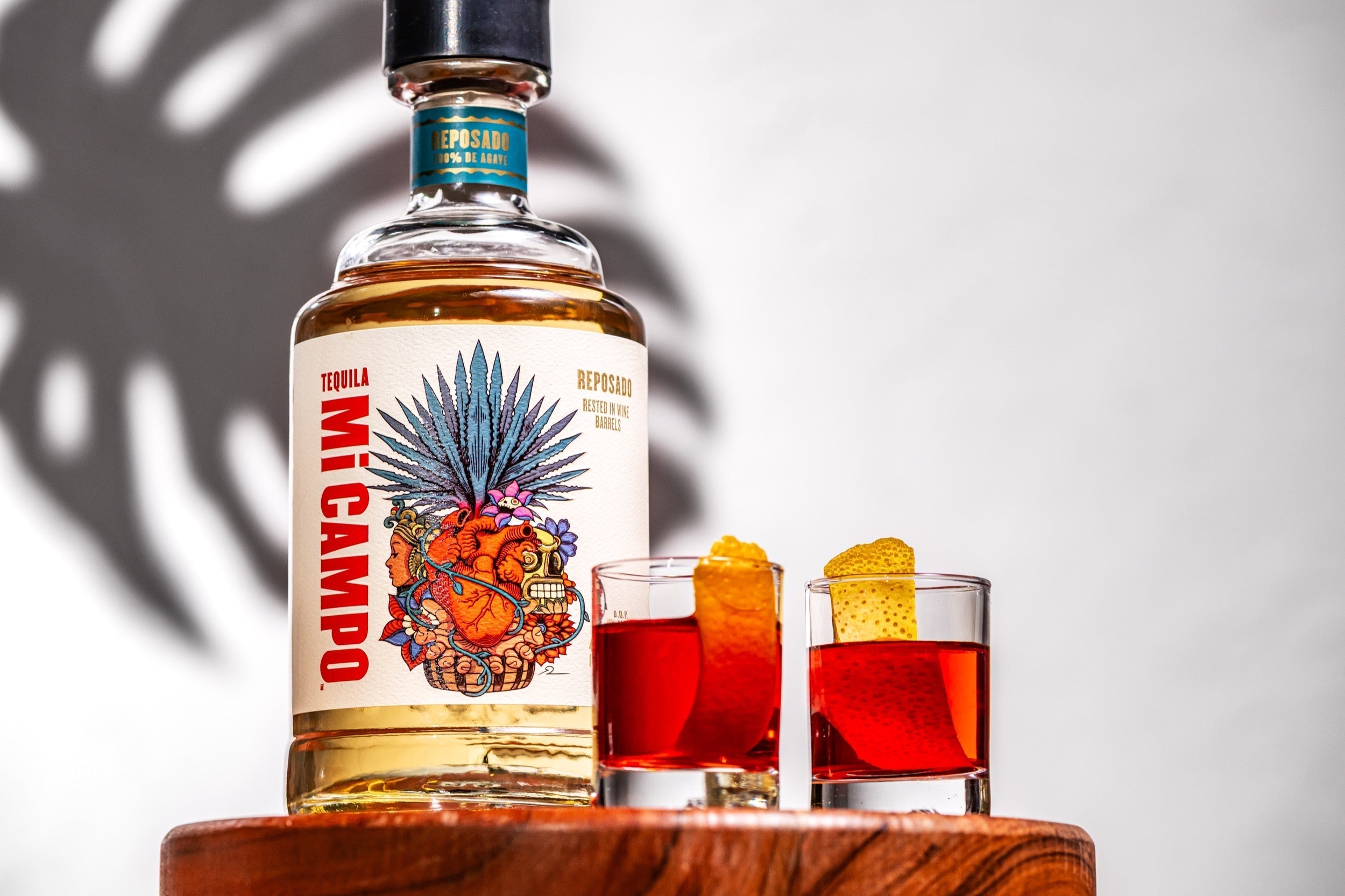 Bottle of Mi Campo Reposado tequila beside two red Rosita shooters garnished with orange peel, set on a wooden surface with soft shadows in the background.