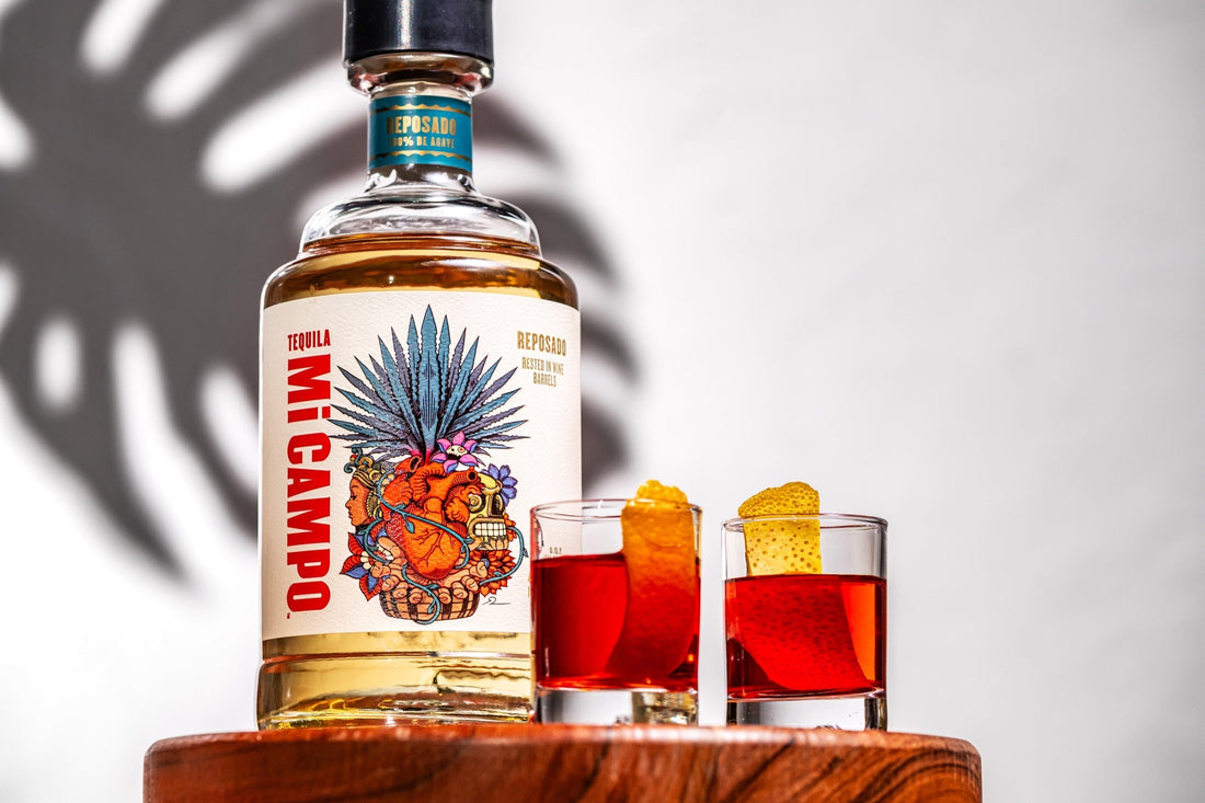 Bottle of Mi Campo Reposado tequila beside two red Rosita shooters garnished with orange peel, set on a wooden surface with soft shadows in the background.