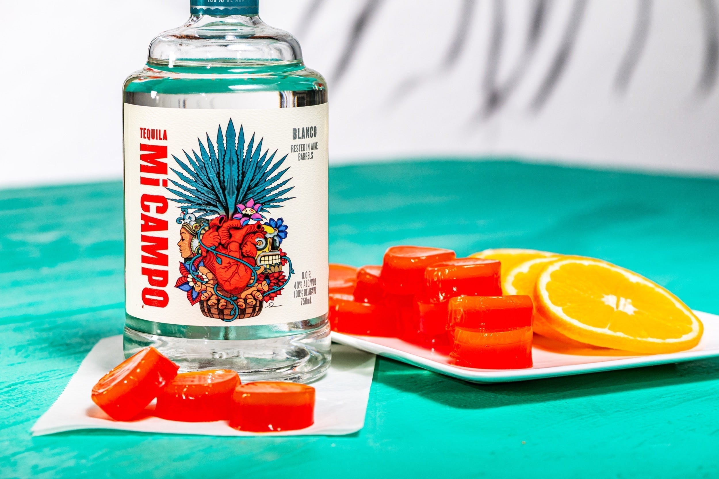 Bottle of Mi Campo Blanco tequila beside tequila sunrise jelly shots and fresh orange slices on a plate.