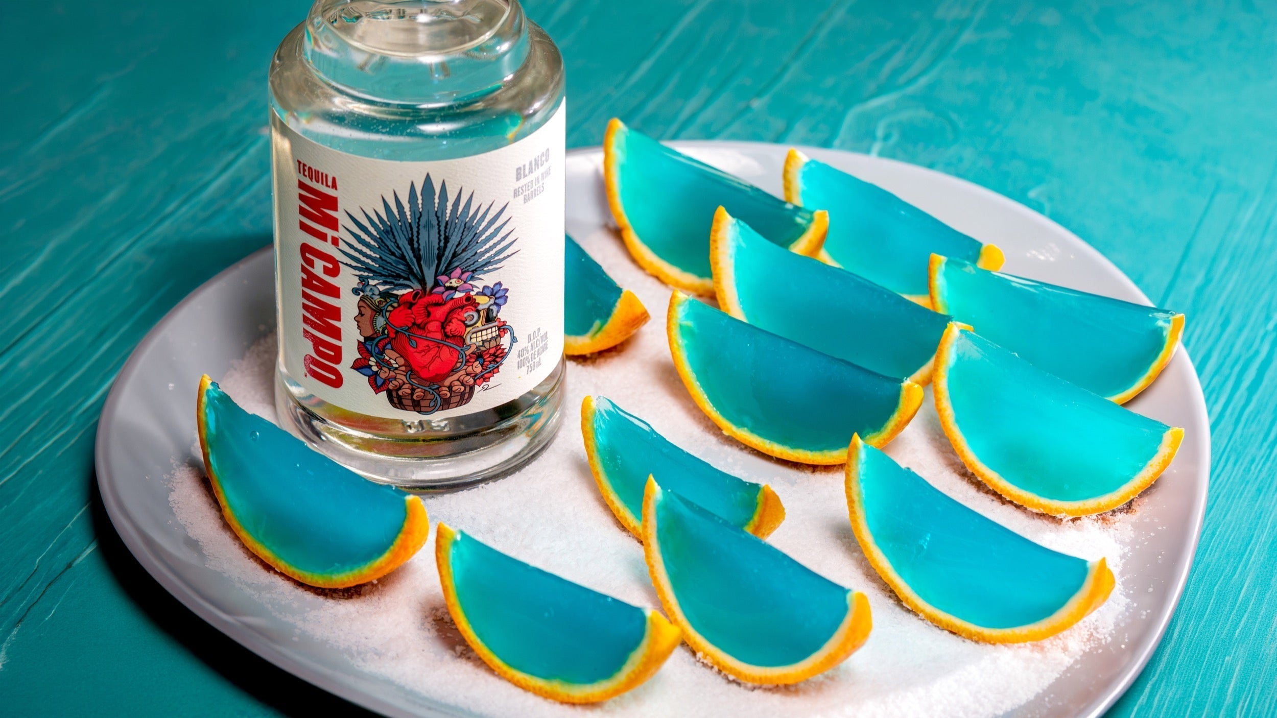A bottle of Mi CAMPO Blanco Tequila is displayed on a white plate covered with coarse salt. Arranged around the bottle are several bright blue margarita jelly shots set inside orange peel wedges, resembling citrus slices.