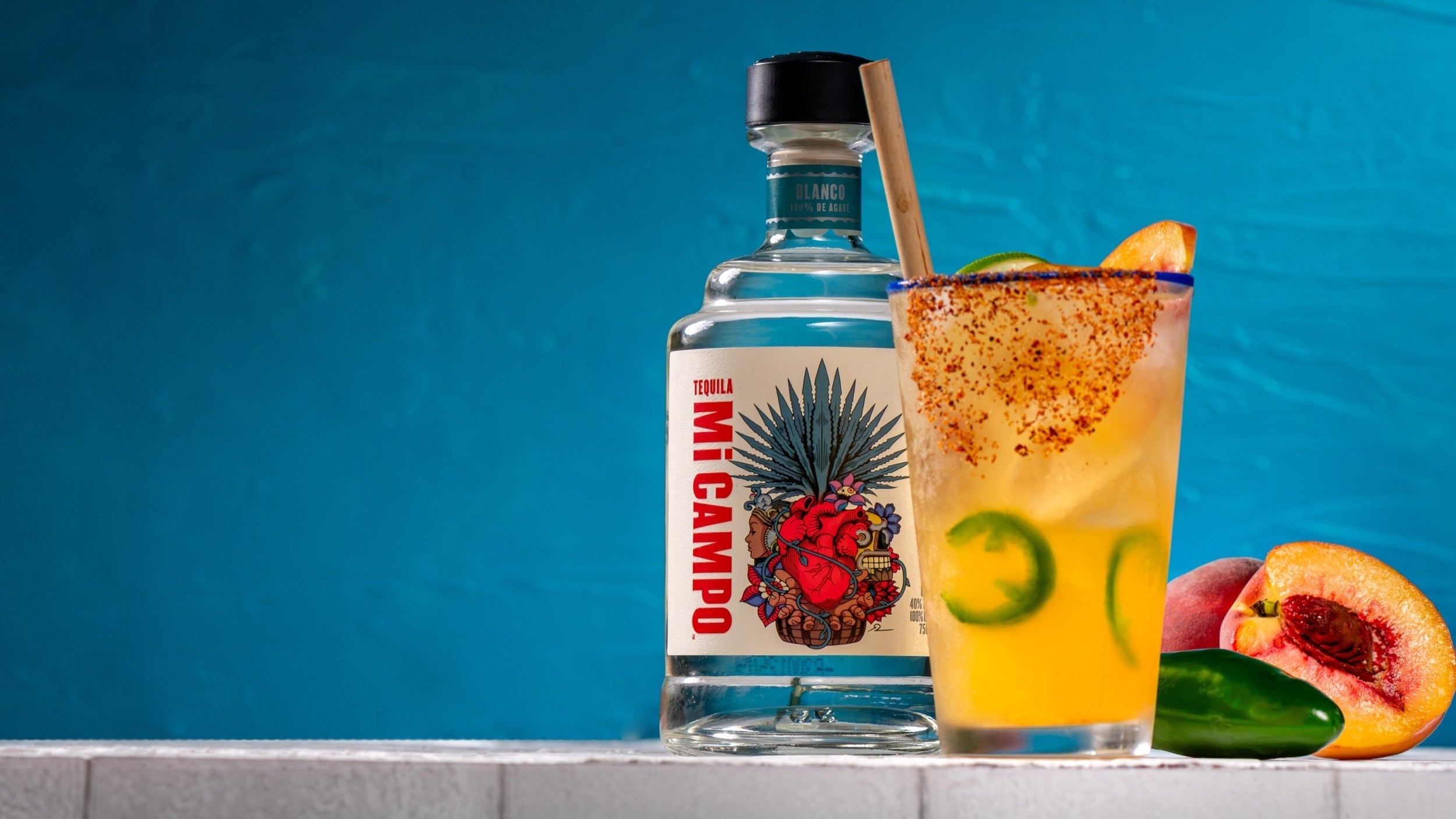 A bottle of Mi CAMPO Blanco Tequila is displayed next to a spicy peach ranch water cocktail served over ice in a clear glass with a chili-seasoned rim.