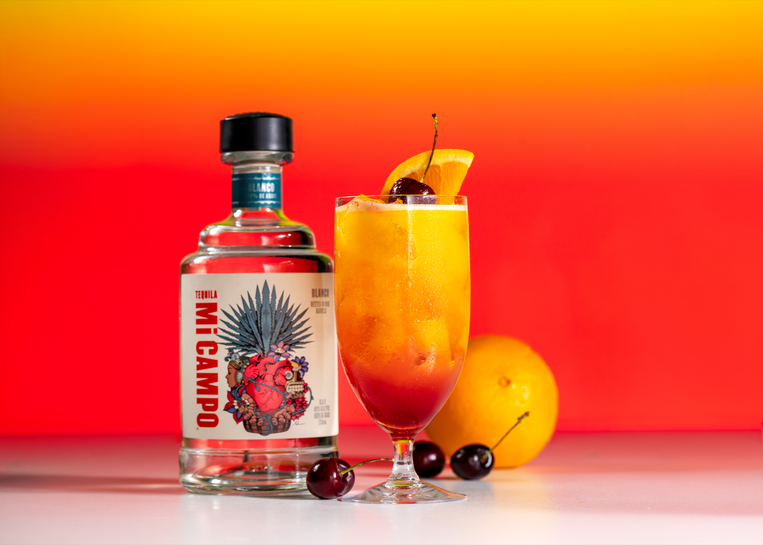 A vibrant image with a yellow to red gradient in the background. On the white surface in front of the gradient sits a bottle of Mi CAMPO Blanco, an orange, and several black cherries surrounding a tequila sunrise cocktail