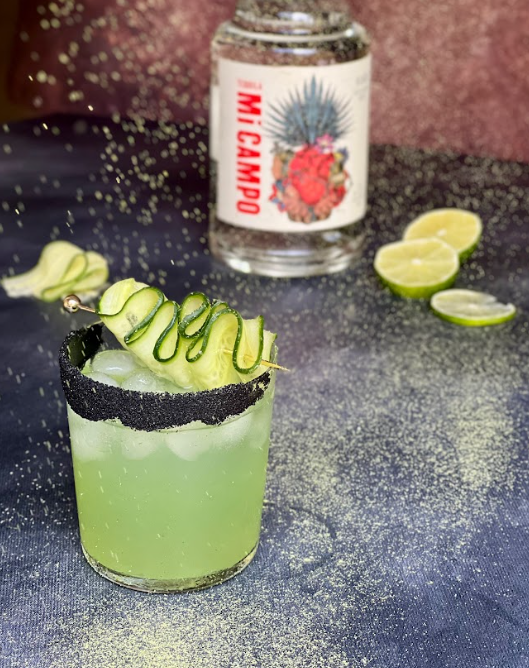 Green cocktail with bottle of Mi Campo in the background and sliced limes