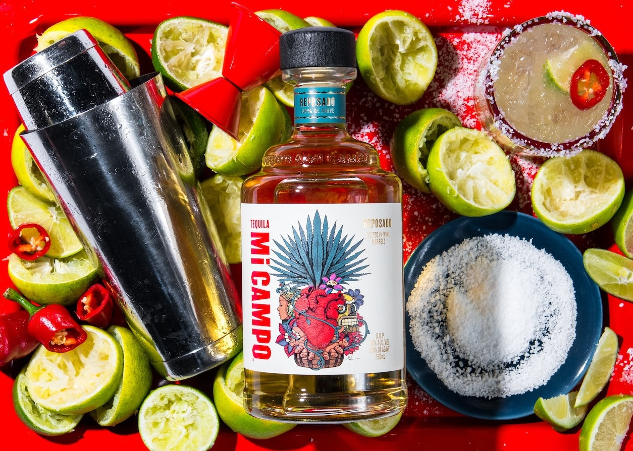 Bottle of Mi Campo Tequila surrounded by limes, salt, and a cocktail shaker on a red background