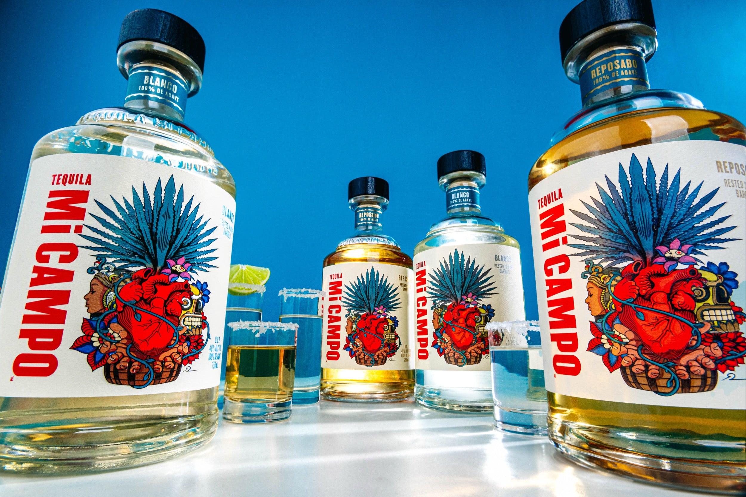 several Mi Campo bottles in both varieties with shot glasses that have salt and lime on the rims taken against a blue background