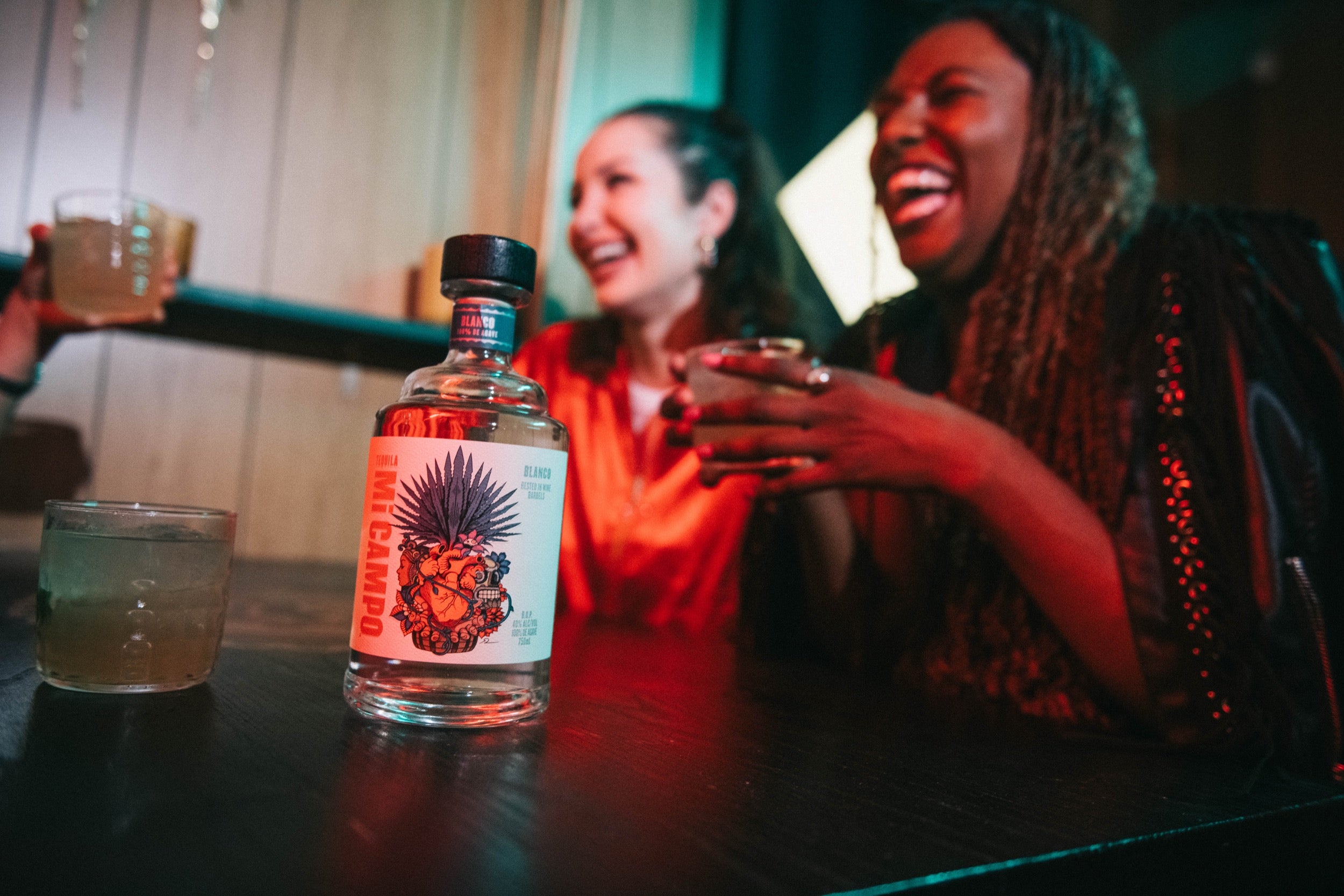 Two people enjoying drinks at a bar with a bottle of alcohol in the foreground.