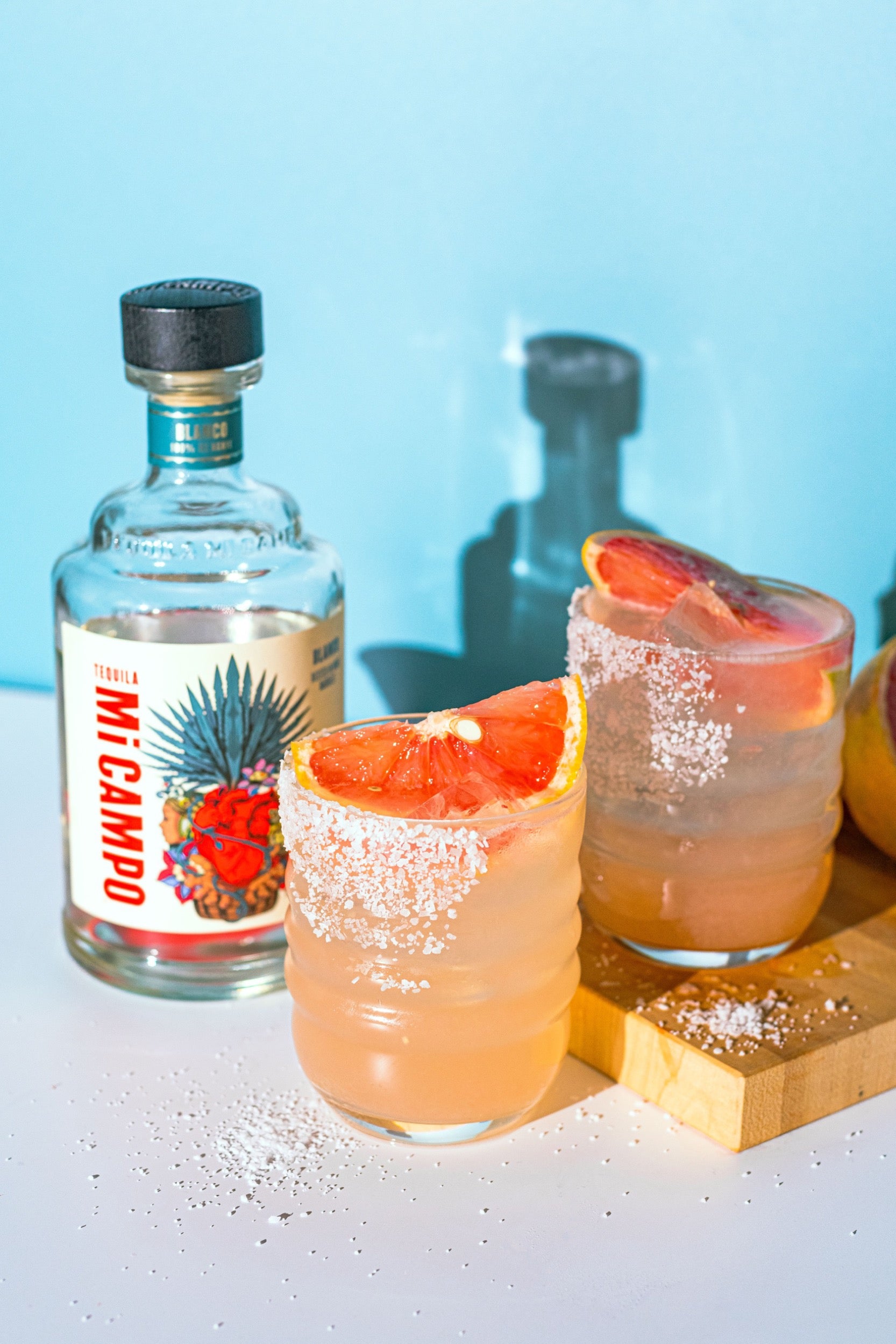 Two glasses of pink grapefruit cocktails with a bottle of Mi Camilo tequila on a light blue background.